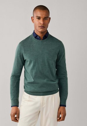 Mann mit kurzen Haaren trägt einen grünen Langarmpullover über einem dunkelblauen Hemd mit Kragen und weißen Hosen und steht vor einem einfarbigen Hintergrund.