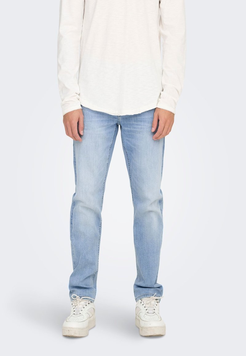 Jeans de mezclilla azul claro con corte recto, combinados con una camiseta de manga larga color crema y zapatillas blancas. Textura suave, diseño casual.