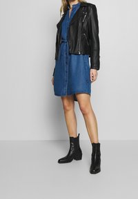 Kvinna iklädd en midilång blå denimklänning med framknappar, en svart läderjacka över axlarna och svarta ankelboots.