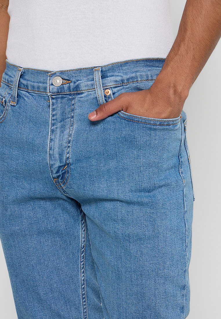 Jean en denim bleu clair avec une coupe ajustée, présentant une poche avant, un bouton en métal et des détails de couture visibles. Le tissu a une texture classique.