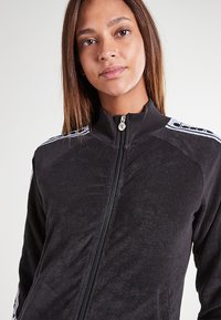 Giacca nera con zip, realizzata in un tessuto testurizzato e con colletto alto. Presenta strisce bianche sulle spalle e una zip in metallo.