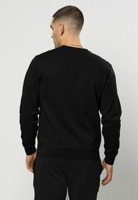 Bărbat cu păr scurt purtând un hanorac simplu negru și pantaloni negri, stând cu fața spre spate pe un fundal neutru.