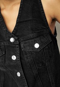 Schwarze Denim-Weste mit tiefem V-Ausschnitt, silbernen Druckknöpfen und Fronttaschen aus strukturiertem Stoff für zusätzliche Details.