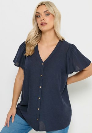 Nainen, jolla on pitkät vaaleat hiukset, pukeutunut tummansiniseen V-pääntielliseen puseroon, jossa on liehukevät hihat ja puiset napit, yhdistettynä vaaleansinisiin farkkuihin.