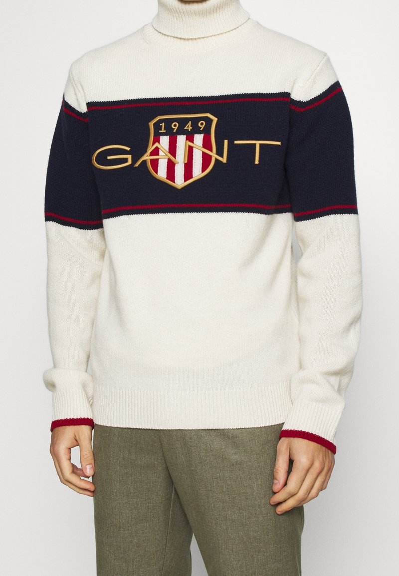Crème col roulé trui met marineblauwe en rode strepen, voorzien van een groot schildlogo met "GANT" en "1949," gebreide textuur, ribbelmanchetten en onderkant.