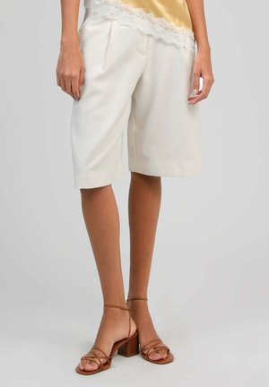 Shorts blancs sur mesure longueur genou portés avec des sandales à talons carrés brunes à brides, sur une personne debout devant un fond uni.