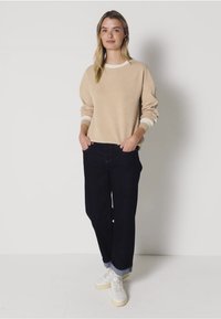 Sudadera beige con puños blancos, combinada con jeans azul oscuro. El modelo lleva zapatillas blancas. Corte simple y relajado, textura lisa.