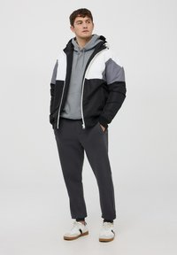 Giacca puffer color block in nero, bianco e grigio, con zip e cappuccio. Indossata sopra un hoodie grigio con pantaloni scuri a gamba affusolata e sneakers.