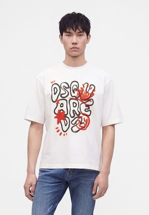 Jeune homme portant un t-shirt blanc à manches courtes avec un texte graffiti abstrait noir et rouge et des formes, associé à un jean bleu, sur fond uni.