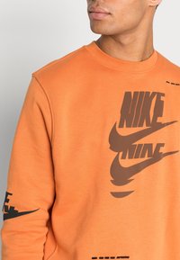 Man som bär en orange Nike-hoodie med stora bruna logotyper på bröstet och en mindre logotyp på ärmen, vänd något mot höger.