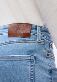 Φως μπλε τζιν από τζιν ύφασμα με καφέ δερμάτινη ετικέτα Calvin Klein στη ζώνη, φορεμένα με λευκό πουκάμισο μαζεμένο μέσα.