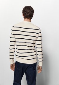 Beige gestreifter Pullover mit marineblauen Streifen, rundem Halsauschnitt und gerippten Bündchen. Kombiniert mit dunklen Jeans.