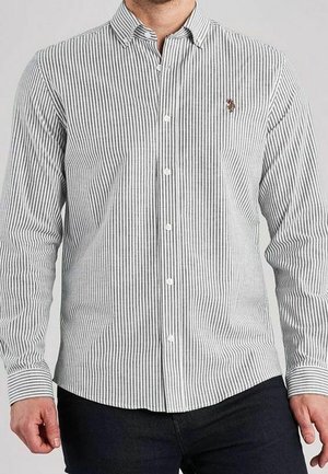 Homme portant une chemise blanche à boutons avec de fines rayures noires verticales et un petit logo brodé sur le côté gauche de la poitrine.