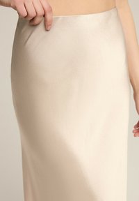 Sukienka midi z satyny w jasnym beżu, gładka tekstura, dopasowany krój, ukazująca rękę delikatnie ciągnącą za pas. Styl minimalistyczny.
