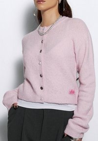 Różowy kardigan zapinany na guziki, wykonany z miękkiego, puszystego materiału, z perłowymi i metalicznymi guzikami, ściągaczami na rękawach oraz metką na dolnym brzegu.