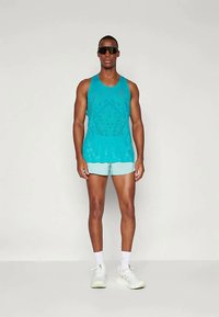 En teal tanktop har et tekstureret design og er parret med lyseblå shorts. Modellen har solbriller og hvide sneakers på.