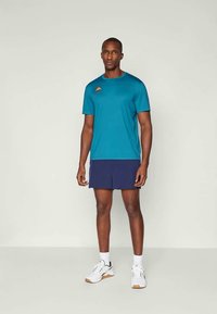 T-shirt atlético turquesa com mangas curtas e logótipo, combinada com calções azul-marinho. Os sapatos são brancos com detalhes cinzentos e cor de goma.