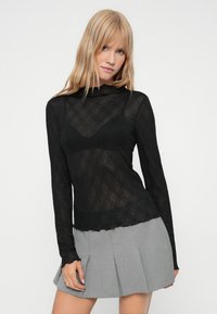 Top din plasă neagră cu mâneci lungi, cu o textură florală, suprapus peste un sutien negru subtil, asortat cu o fustă plisată gri.