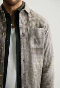 Camicia a quadri in tonalità calde, con taschino, design a bottoni, indossata sopra una camicia verde scuro con texture e una maglietta bianca sotto.