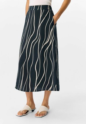 Nainen, joka on pukeutunut mustaan midi-hameeseen, jossa on vaaleanbeigejä aaltoilevia raitoja ja taskuja, yhdistettynä vaaleanbeigeihin avonaisiin korkokenkiin.