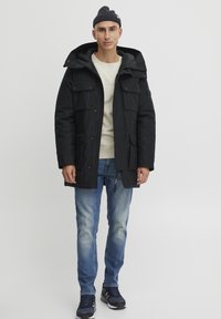 Sort isoleret parka med hætte, flere frontlommer, en lynlås og trykknaplukninger, båret over en lys sweater og blå jeans.