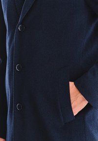 Granatowy wełniany płaszcz z klapami, pięcioma guzikami i kieszenią boczną. Tekstura jest gładka, a kształt dopasowany.