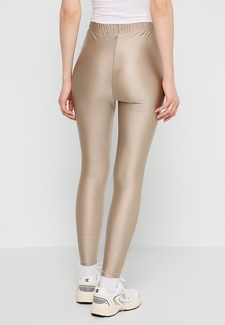 Beżowe legginsy z wysokim stanem, błyszczące, wykonane z elastycznej tkaniny, z elastycznym pasem i dopasowanym krojem. Noszone z białymi trampkami.