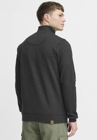 Pull gris foncé avec un col haut, des poignets et un ourlet côtelés. Présente une petite étiquette logo à l'arrière et une texture de tissu lisse.