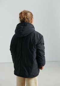 Must sulgepõhise jopega, kapuutsiga, tikandmustriga ja lõdva lõikega. Lihtne disain elastsete varruka- ja allääristega, kantud beežide pükste kohal.