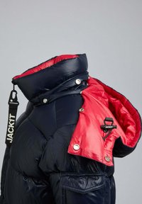 Marinblå isolerad pufferjacka med röd insida, multifunktionell huva, glänsande finish, tryckknappar och ett märke med "JACKIT" logotyp.