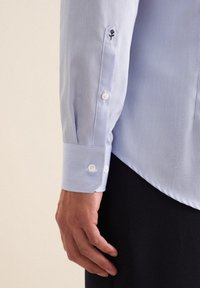 Lichtblauw gestreept shirt met knoopmanchetten; bevat een klein zwart logo op de mouw; de stof lijkt glad en heeft een lichte textuur.