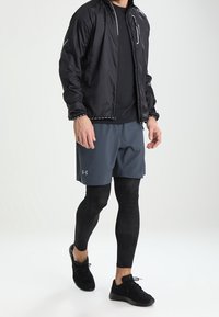 Veste légère noire avec accents réfléchissants, t-shirt noir à col rond, short gris, leggings texturés noirs et chaussures de sport noires.
