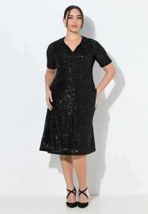 Kvinde står iført en knælængde sort pailletkjole med korte ærmer og sorte stropper hæle mod en hvid baggrund.