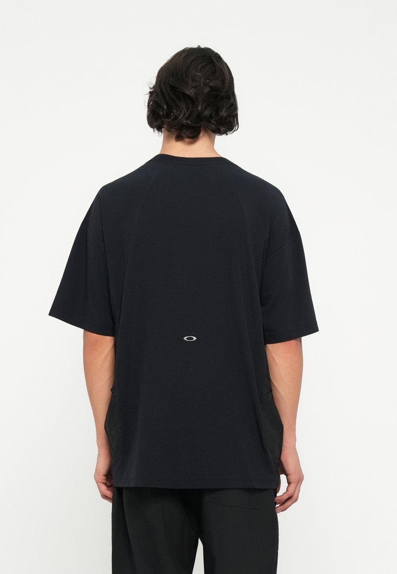 Camiseta negra de gran tamaño con cuello redondo, mangas cortas y detalles de tejido en contraste en los lados, con un detalle de logotipo cerca del dobladillo.