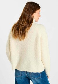 Vrouw met lang bruin haar, gekleed in een crèmekleurige gebreide trui en blauwe jeans, met de zijkant naar de camera gericht tegen een eenvoudige witte achtergrond.