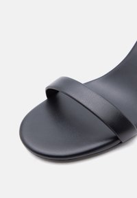 Κοντινό πλάνο ενός μαύρου σανδαλιού με ανοιχτά δάχτυλα και λουράκι πάνω σε μία λείος σόλα με μινιμαλιστικό σχέδιο και ματ υφή.