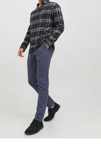 Uomo che indossa una camicia a scacchi scura con bottoni, pantaloni blu slim-fit e scarpe nere stringate, in piedi con una mano in tasca.
