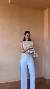 Top beige a maglia con punti aperti e lacci laterali, abbinato a pantaloni in lino bianchi. Tiene in mano un tessuto a righe verde chiaro con un'etichetta.