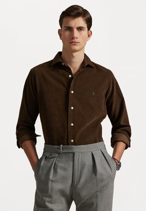 Camisa de cotelê castanha com um logótipo verde, apresentando um colarinho clássico e fecho de botão, combinada com calças plissadas cinzentas de cintura alta.
