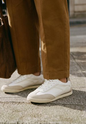 Hvite sneakers med beige accenter båret med brune bukser med vide ben på en brolagt udendørs overflade ved siden af en brun ruskindstaske.