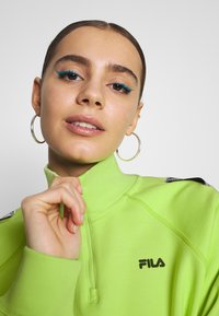 Felpa verde con zip e colletto alto, caratterizzata da un logo nero sul petto e un tessuto texturizzato. Orecchini a cerchio grandi e argentati ben visibili.