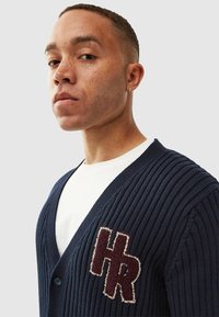 Marinblå ribbad cardigan med v-ringad hals, med en texturerad vinröd "HR"-logotyp. Knäppning med knappar och mjukt stickat material.