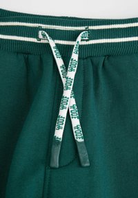 Zielone dresy mają żebrowaną talię z białymi paskami oraz sznurek do regulacji z nadrukowanym tekstem. Materiał jest miękki i teksturowany.