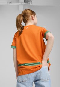 Oranžové tričko s krátkým rukávem a zeleným lemem, s texturovanými vzory na rukávech a kulatým výstřihem, stylizované s světle modrými džíny.