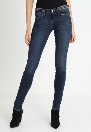 Kobieta w ciemnoniebieskich obcisłych dżinsach z niskim stanem i czarnych botkach na obcasie, stojąca na białym tle.