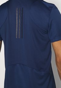 Muž oblečený v námořnickém modrém sportovním tričku se třemi vertikálními černými pruhy vzadu, zobrazený od poloviny zad po paži.