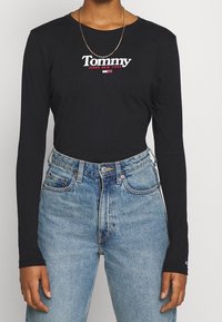 Svart långärmad tröja med vitt och rött "Tommy Jeans New York"-logotyp, matchad med högmidjade ljusblå jeans.