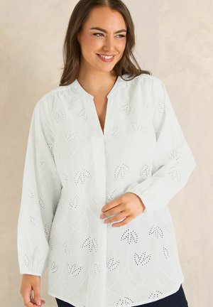 Smilende kvinde iført en hvid langærmet bluse med hulmønster og bølget udskæring mod en neutral baggrund.