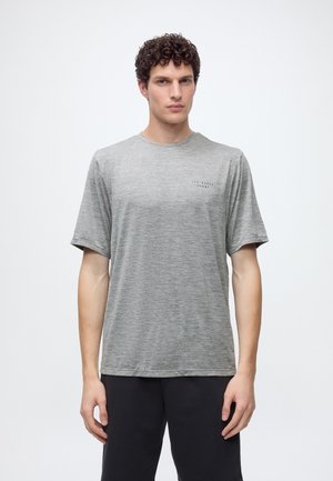 Hombre con cabello rizado que lleva una camiseta gris de manga corta Ted Baker Sport y pantalones negros, de pie frente a un fondo blanco liso.