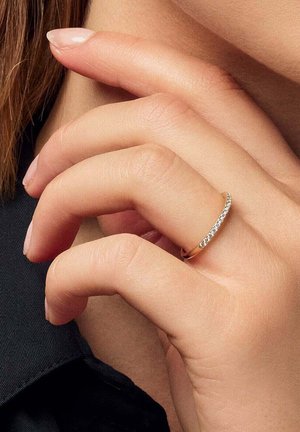 Hånd med naturlige negle iført en tynd guldring med små diamanter, der rører ved ansigtet nær hagen, mod en mørk stofbaggrund.